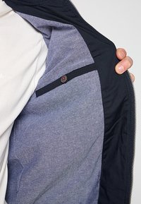 Chaqueta con cremallera de tela exterior azul marino. En el interior presenta un forro gris claro y un bolsillo de tela azul con cierre de botón.