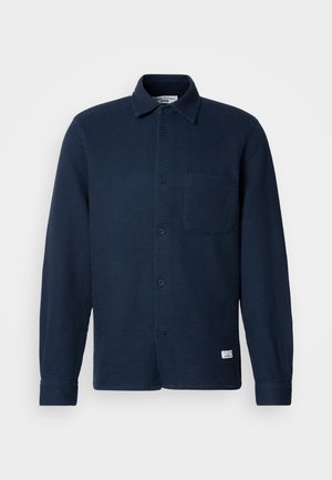 Tengerészkék gombos ing, texturált gofris anyagból, hosszú ujjú, egyetlen mellzsebbel és címkével az alsó szegélyen.