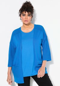 Blå lätt cardigan med nätstruktur på ärmarna, öppen fram, och två framfickor, lagd över en matchande rundhalsad topp.