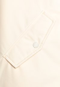 Tessuto beige con una superficie liscia presenta una tasca rettangolare con cuciture a contrasto e un logo circolare con testo.