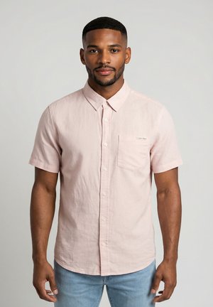 Uomo con capelli corti e barba curata che indossa una camicia rosa chiaro a maniche corte con bottoni e jeans azzurri su uno sfondo uniforme.