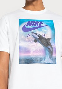 Vit bomulls-T-shirt med en färgglad grafisk bild av en hoppande späckhuggare mot en blå och lila bakgrund med "NIKE AIR" text ovanför.
