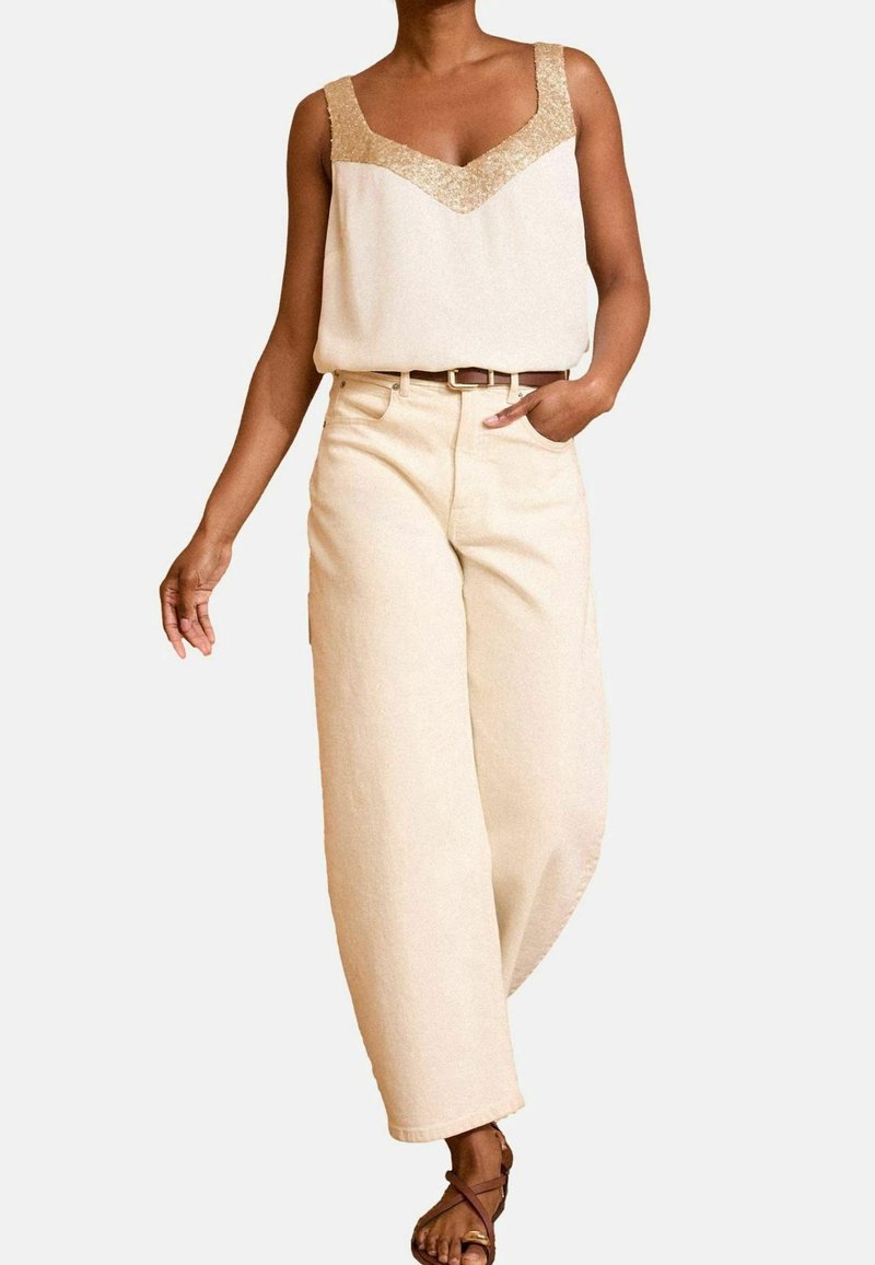Béžový top bez rukávů se zlatými strukturovanými ramínky, v kombinaci s béžovými kalhotami s vysokým pasem a širokými nohavicemi, doplněný hnědým opaskem a sandály na řemínky.
