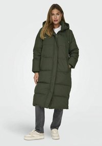 Femme portant un long manteau matelassé vert foncé avec capuche, associé à un jean foncé et des baskets blanches, se tenant devant un fond uni.