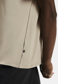 Béžové športové tričko s okrúhlym lemom, obsahujúce bočný rozparok a čiernu nášivku. Hladká textúra a minimalistický dizajn.