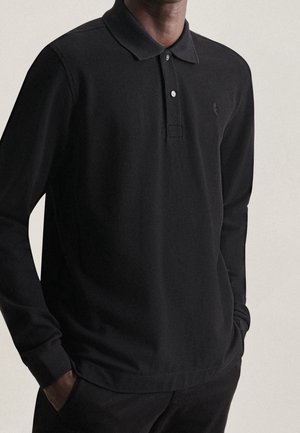 Homme portant un polo à manches longues noir avec col boutonné, mains dans les poches, sur un fond uni clair.