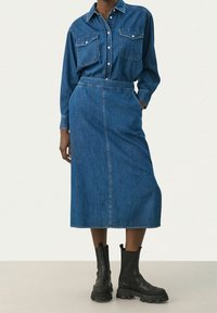 Žena v zapínané džínové košili, kterou má zastrčenou do odpovídající džínové midi skorty s kapsami, doplněná černými robustními kotníkovými botami.