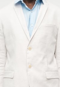 Φωτεινό γαλάζιο πουκάμισο κάτω από ένα λευκό σακάκι με δύο κουμπιά κλειστά και τρεις τσέπες, φωτογραφημένο από τον λαιμό μέχρι τη μέση.