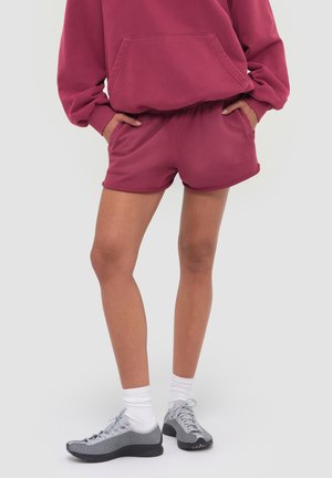 Person iført mørkerosa sweatshirt og matchende shorts med hænderne i lommerne, hvide sokker og grå sports sko.