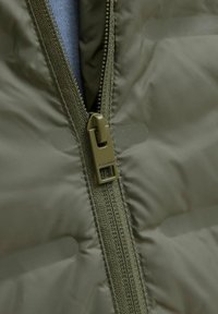 Detailní záběr na olivově zelený zip bundy, částečně rozepnutý, zobrazující strukturu látky a jezdec se značkovým rytím.