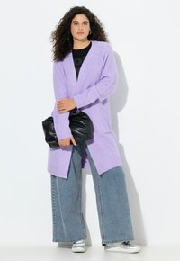 Lawendowy prążkowany długi kardigan z otwartym przodem, noszony na czarnej bluzce, zestawiony z szerokimi niebieskimi dżinsami i srebrnymi trampkami, trzymający czarną torbę.