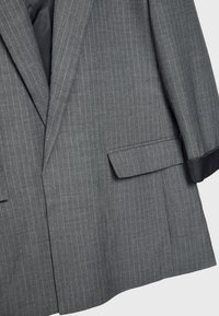 Blazer gris con rayas finas, solapas con muesca, un bolsillo frontal sencillo y forro liso; la tela parece texturizada y confeccionada para un ajuste formal.