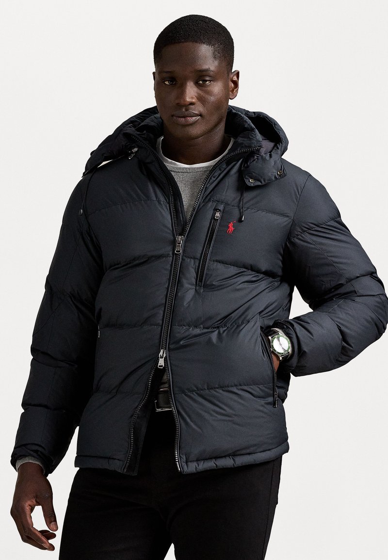 Puffer jacket in dark navy, featuring a hood, zippered front, and red logo accent. Textured fabric with a quilted design and two side pockets.
