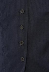 Ρούχο από μπλε σκούρο ύφασμα με ριμπ υφή, με πέντε μαύρα στρογγυλά κουμπιά κατά μήκος μιας κεντρικής πατιλέτας, με λεία υφή και κάθετες γραμμές.