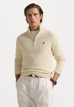Cream-colored zip-up sweater with a ribbed hem and cuffs, featuring a small, dark logo on the left chest. Worn over a light shirt.
