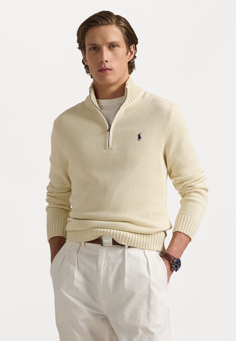 Sweat à zip couleur crème avec un ourlet et des poignets côtelés, présentant un petit logo sombre sur la poitrine gauche. Porté par-dessus une chemise claire.