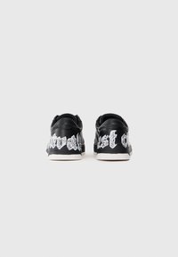 Mustad mustad sneakers valge gooti tekstiga, mis on prinditud ümber kanna ja valged tallad, nähtavad tagantpoolt valgel taustal.