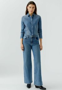 Lichtblauw denim overhemd met drukknoopsluitingen, gecombineerd met wijde lichtblauwe jeans. Zwarte enkellaarsjes maken de outfit compleet.