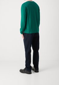 Zielona koszula z długim rękawem, z okrągłym dekoltem, w połączeniu z ciemnymi spodniami i czarnymi butami. Tkanina wygląda na gładką, z luźnym krojem.