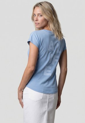 Blonde Frau trägt ein hellblaues T-Shirt mit vertikalem "ALIFE"-Schriftzug auf dem Rücken, kombiniert mit einem weißen Rock, schaut über ihre Schulter.