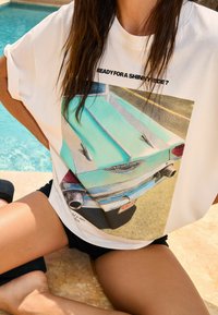 T-shirt blanc avec un graphisme de voiture vintage en turquoise. Coupe décontractée avec des manches courtes et un col rond. Short noir visible en dessous.