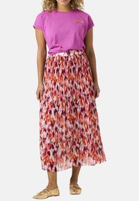 Roze kortarm T-shirt met een klein logo, gecombineerd met een lange, geplisseerde rok in levendige tinten van oranje, roze en wit. Gouden flats.