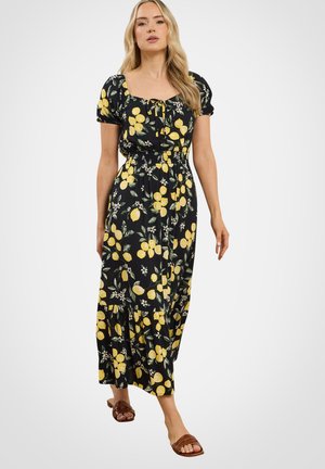 Vrouw draagt een zwarte midi-jurk met gele citroen- en groene bladdruk, pofmouwen en bruine sandalen met open tenen, lopend naar voren.