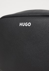 Sort teksturert lærveske med en glatt topp; har en hvit "HUGO"-logo på forsiden. Strømlinjeformet, kompakt design med subtile detaljer.