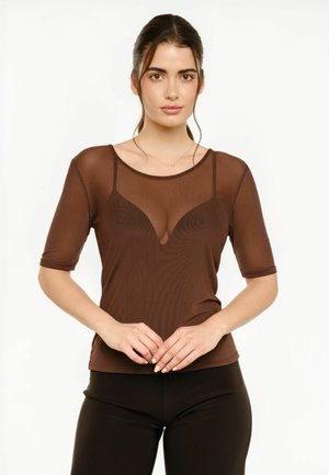 Frau mit dunklem zurückgebundenem Haar, trägt ein durchsichtiges braunes Top über einem braunen Bustier, kombiniert mit schwarzen Hosen, stehend vor weißem Hintergrund.