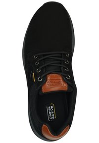 Zapatilla deportiva negra con un upper de textura suave, cordones negros y un acento de cuero marrón cerca de la lengua. Incluye detalles del logo.