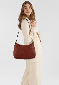 Donna in completo crema che porta una grande borsa a tracolla in pelle marrone, sorridendo e guardando di lato contro uno sfondo bianco semplice.