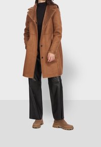 Manteau marron en mélange de laine avec un col en fausse fourrure, doté d'une fermeture à boutons et de poches latérales, porté avec un pantalon en cuir noir et des chaussures beige épaisses.