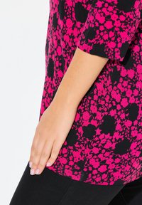 Schwarzblumiges Oberteil mit einem lebhaften pinken und schwarzen Blumenmuster, kurzen Ärmeln, weichem Stoff und gerundetem Saum. Nahaufnahme einer Hand auf dem Stoff.