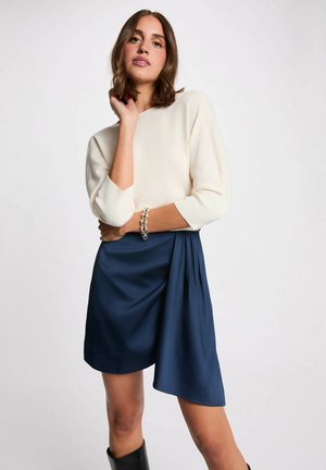 Donna che indossa una maglia a maniche lunghe color crema, gonna asimmetrica blu navy, bracciale a catena argento e stivali neri, in piedi davanti a uno sfondo chiaro e uniforme.