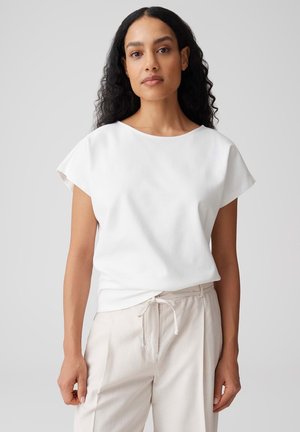 Vrouw met lang krullend haar, gekleed in een wit T-shirt met korte mouwen en lichtbeige broek met een taillekoord, staat tegen een effen achtergrond.