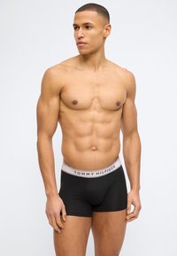 Μυώδης άντρας όρθιος, φορώντας μαύρα μποξεράκια Tommy Hilfiger με λευκή λείανση με λογότυπο, κοιτάζει προς τα αριστερά του σε απλό φόντο.