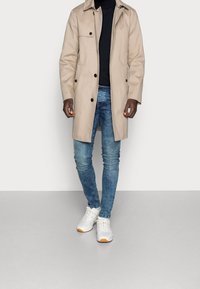Trench-coat beige avec col et boutons, associé à un jean bleu foncé et des baskets blanches. Texture lisse, coupe ajustée et design moderne.