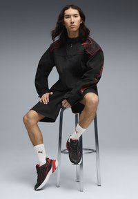 Veste de sport noire avec des accents rouges, associée à un short noir. Baskets noires et rouges, chaussettes blanches avec logo. Assis sur un tabouret en métal.
