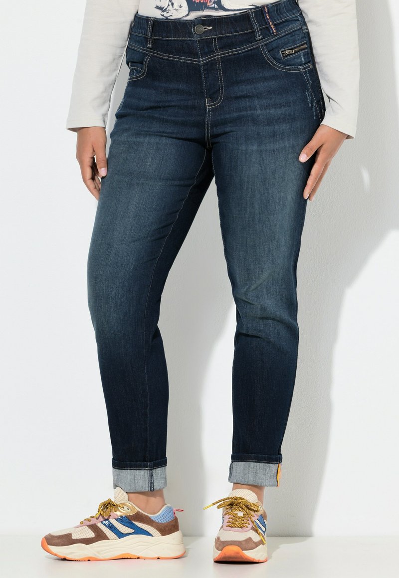 Tmavě modré džíny s vysokým pasem, ohrnutými nohavicemi a kontrastním prošíváním. Nosené s barevnými teniskami, které mají různé textury a barvy.