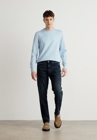 Ανοιχτό μπλε πουλόβερ με μακριά μανίκια, σκούρα τζιν και μπεζ μπότες. Το πουλόβερ έχει στρογγυλή λαιμόκοψη και ένα μικρό λογότυπο στο στήθος.