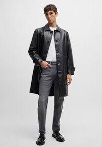 Sort læder trench coat med knaplukning foran, lange ærmer og manchet detaljer, parret med grå jeans og sorte sko.