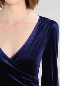 Námořnická modrá sametová halenka s dlouhými rukávy a výstřihem do V. Má jemnou texturu a zavinovací design, který se shromažďuje v pase.