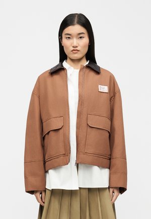 Γυναίκα με μακριά ίσια μαλλιά φοράει καφέ μπουφάν με μεγάλες τσέπες πάνω από λευκό πουκάμισο και πλισέ μπεζ φούστα, στέκεται μπροστά σε απλό φόντο.