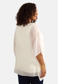 Lättviktig, genomskinlig vit blus med svaghängande ärmar, en avslappnad passform och rundad kant. Bärs över mörk denim med subtil struktur.