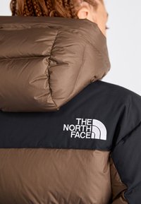 Smešani smeđi i crni vodootporni kaput s visokim ovratnikom. Sadrži bijeli logo The North Face s lijeve strane. Glatka, teksturirana tkanina s blagim sjajem.