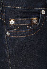 Mörkblå denimtyg med blekt mönster och gula sömmar; har en liten framficka med metallnitar och triangulära detaljer.