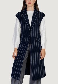 Námořnický pruhovaný dlouhý vest s klopami, dvěma předními kapsami a vycpávkami na ramenech, nošený přes bílý svetr s pleteným vzorem.