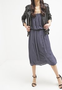 Grå lång klänning med en flödande silhuett, lager-på-lager under en svart läderbikerjacka. Har en axelbandslös halsringning och mjuk textur. Svarta sandaler med klack.