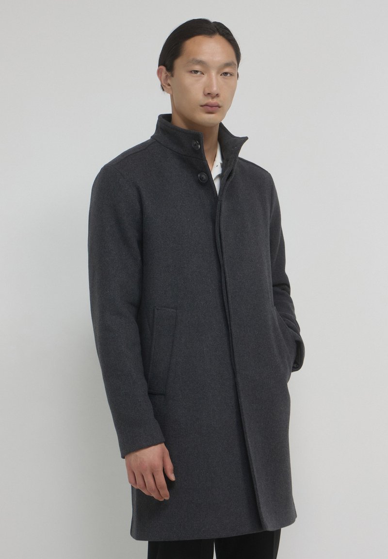 Uomo con capelli scuri che indossa un cappotto grigio scuro con bottoni e tasche laterali, in piedi contro uno sfondo grigio chiaro uniforme.