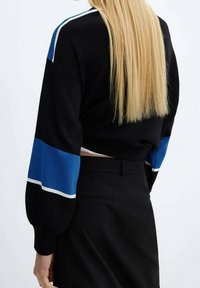 Pull court noir avec accents rayés bleus et blancs sur les manches, associé à un pantalon noir taille haute. Texture lisse, coupe décontractée.